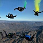 Dynamic aerial shot of skydivers performing stunts with colorful smoke trails over mountainous terrain.
