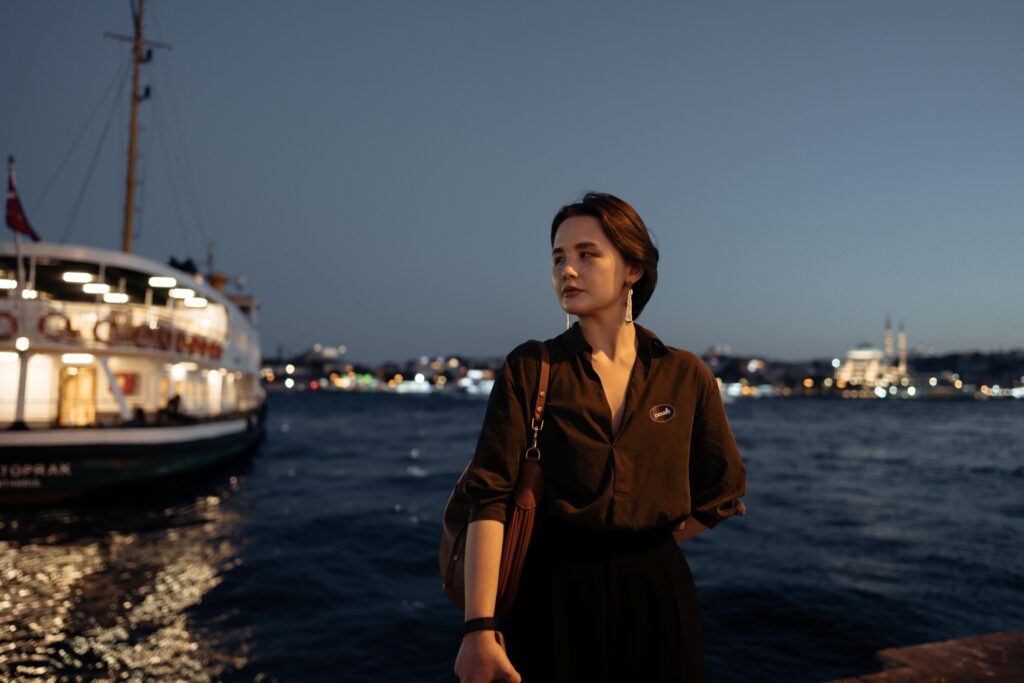 Woman standing by the Bosphorus in Istanbul at twilight, with city lights and a ferry in the background.